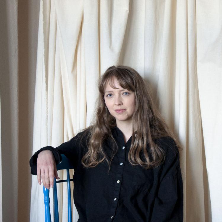 Porträtt på Emilia Elfvik. Emilia har svart skjorta och står framför ett beige drapperi