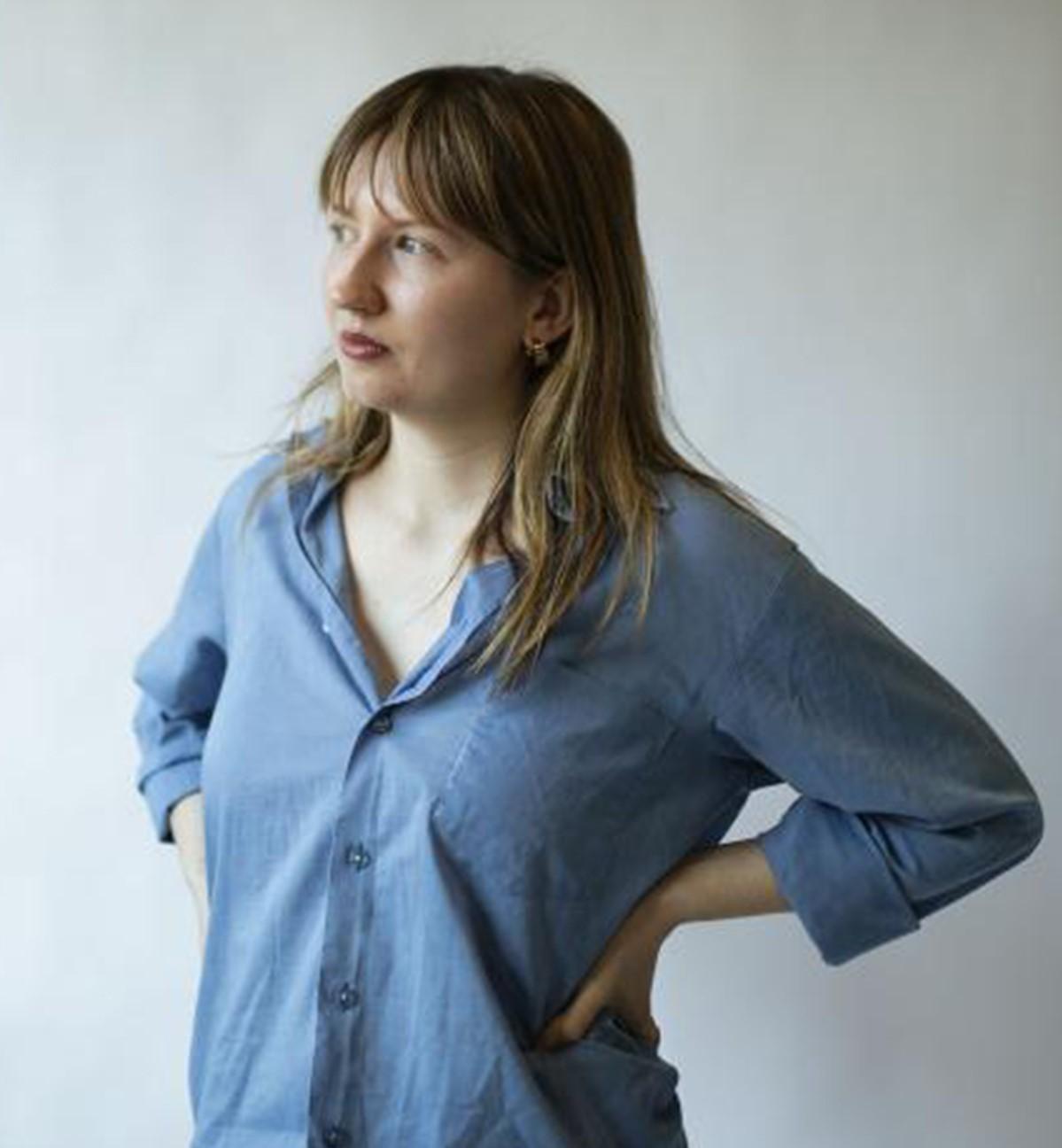 Foto på Simone Norberg i en inomhus studiomiljö. Simone har på sig en ljusblå skjorta och jeans. Simones blonda hår hänger ned över axlarna medans hon tittar bort mot vänster.