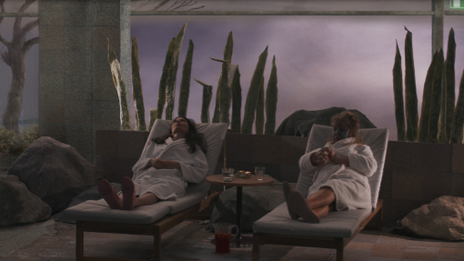 Stillbild på två kvinnor som ligger ner på solstolar i en inomhus spa-miljö. Den ena kvinnan lutar sig tillbaka och ser ut att försöka koppla av. Den andra kollar på hennes klocka.